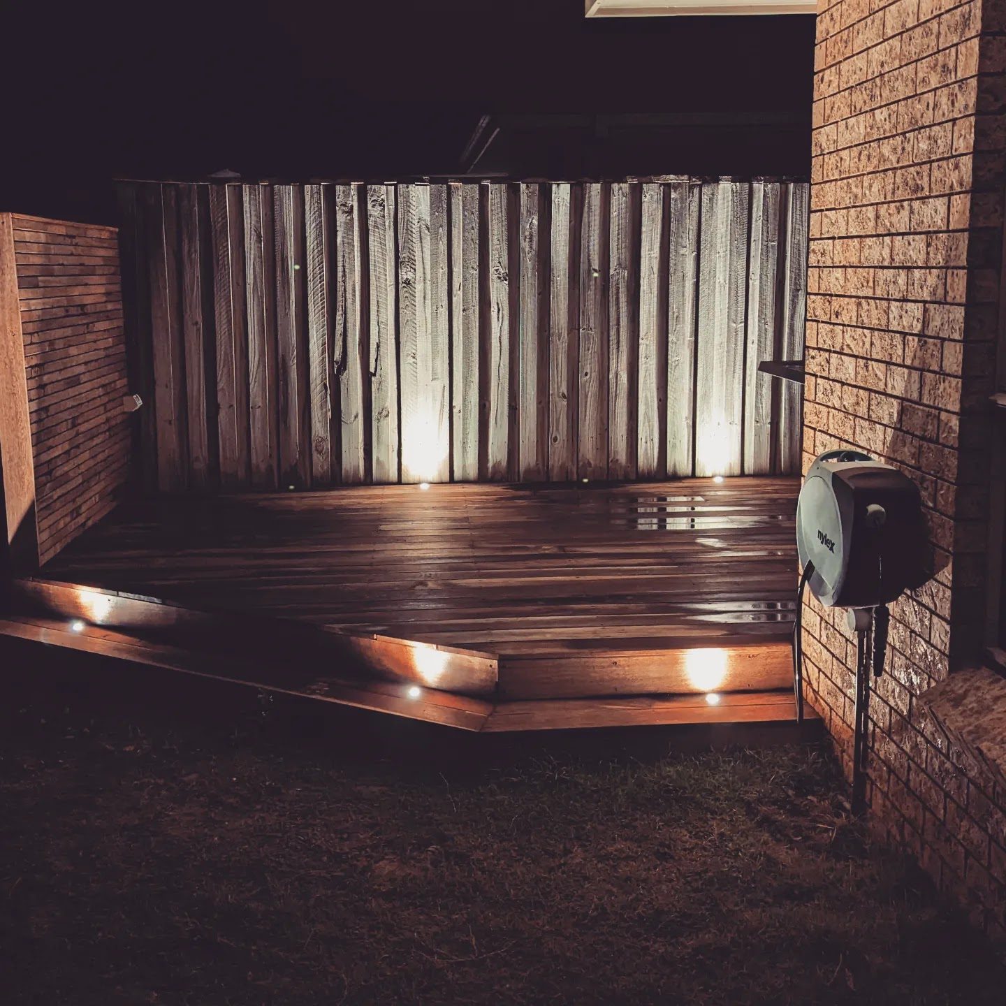 A timber deck at night, lit by recessed lights in the boards and uplights on the fence